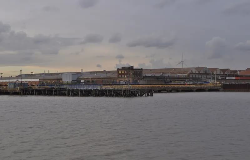 A dock with several industrial buildings under a cloudy sky, with a dark body of water in the foreground.