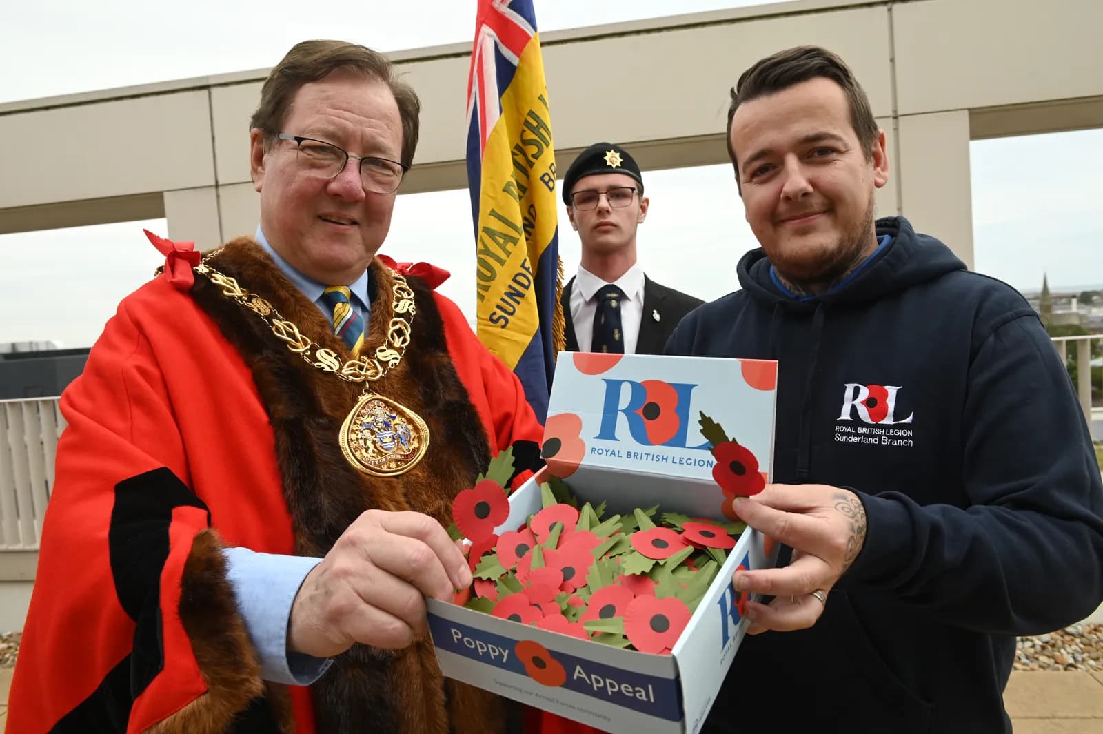 Two men, one in a ceremonial red robe and gold chain, and another in a blue hoodie, hold a box of paper poppies for the Royal British Legion Poppy Appeal.