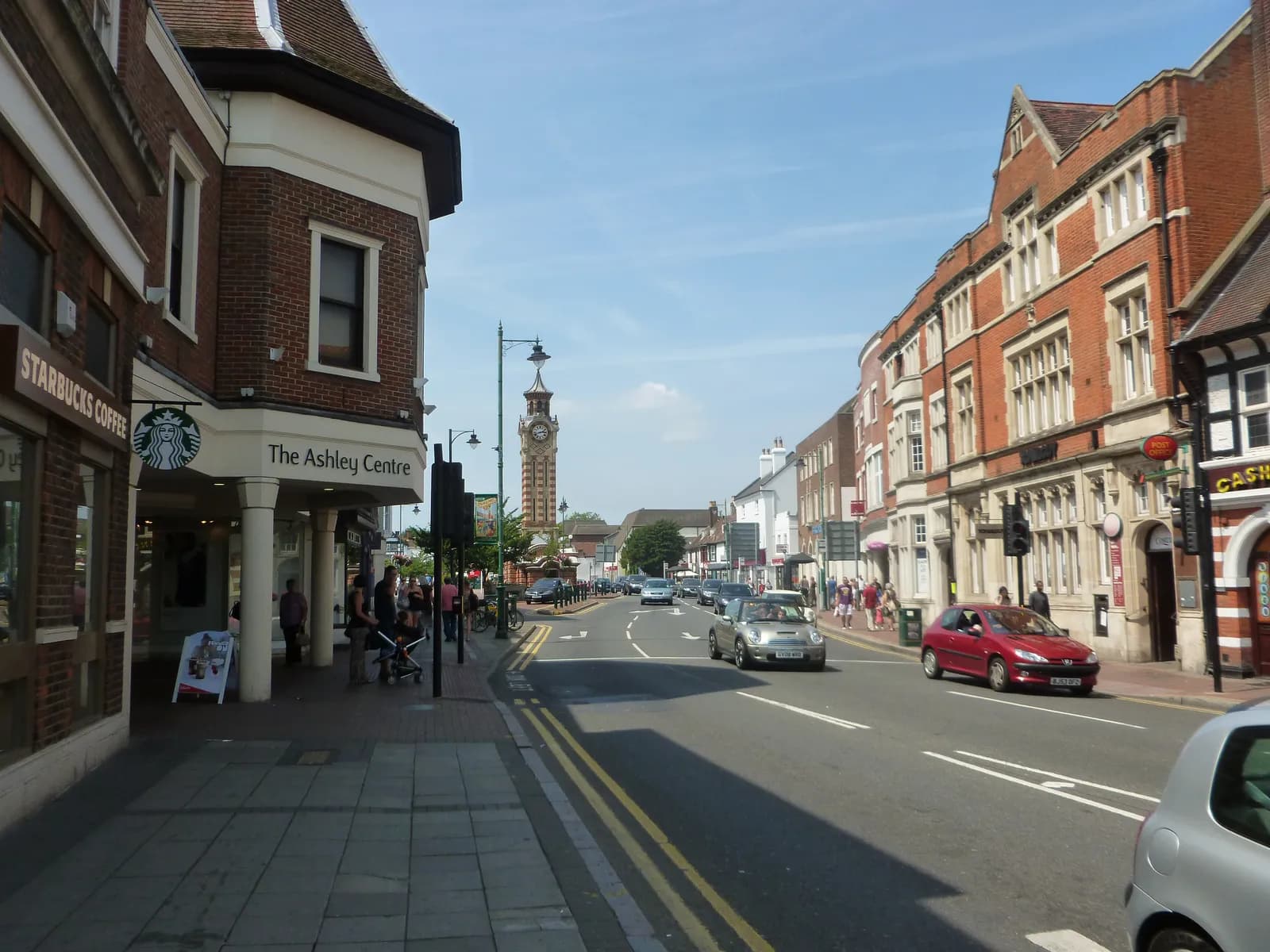 A clock tower stands tall in the distance over a busy street lined with brick buildings, cars, and pedestrians on a sunny day.