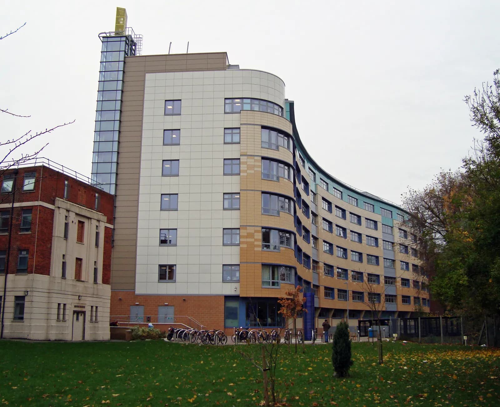 A modern building with a curved facade composed of varying shades of tan, blue, and white panels, stands next to an older red brick building, both fronted by a green lawn with scattered autumn leaves and bicycle racks.