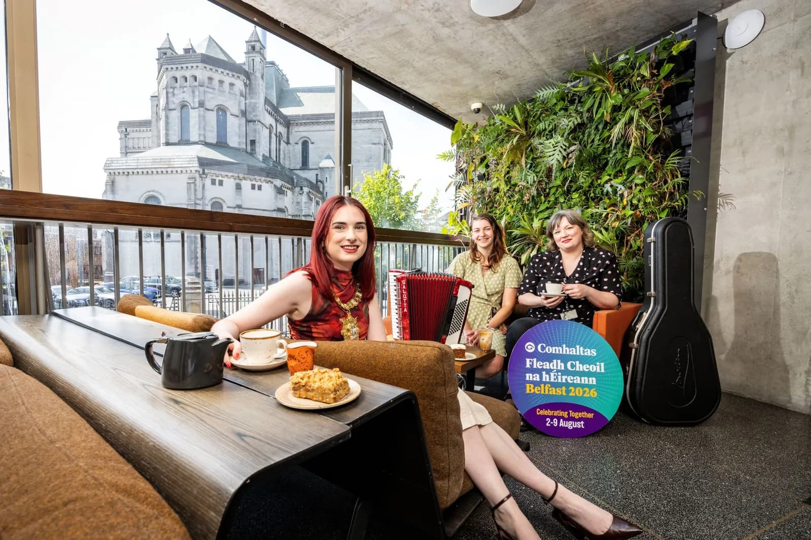 Three women gathered in a cafe, one with red hair holding a teacup, one with an accordion, and one sipping coffee, with a large building visible outside a window and a 'Comhaltas Fleadh Cheoil na hÉireann Belfast 2026' sign.