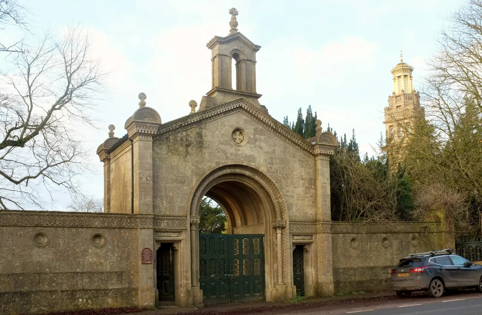 A stone archway with a heavy green wooden gate stands between two stone walls, leading to a tall, neoclassical-style tower visible in the background.
