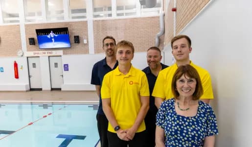 A group of five people, three men in yellow shirts, one man in a black shirt, and one woman in a blue patterned top, stand smiling next to a swimming pool.