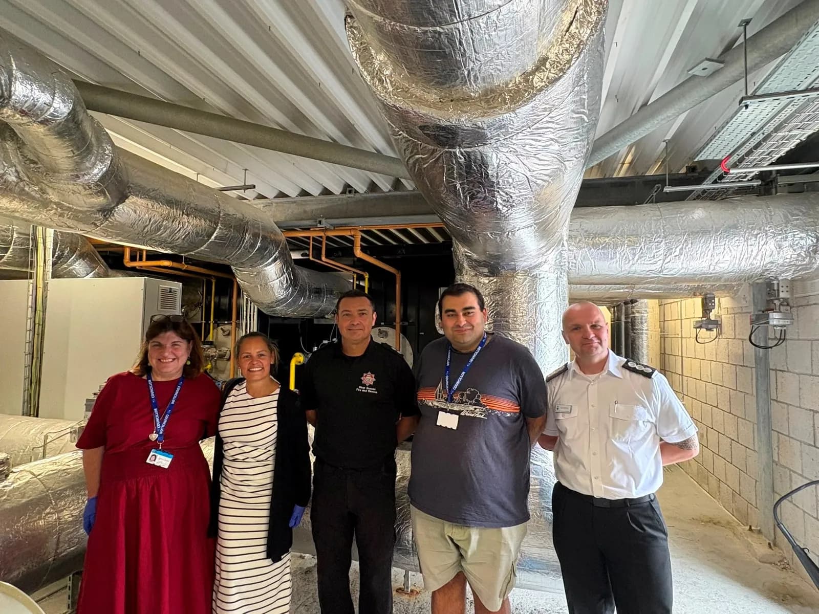 Five adults stand smiling in what appears to be an industrial room with large, insulated pipes running overhead.