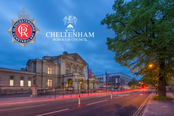 A nighttime street scene shows a classical building with illuminated windows, a tree, and a road with red light trails from moving vehicles, under a dark blue sky featuring the logos for Gloucestershire Constabulary and Cheltenham Borough Council.
