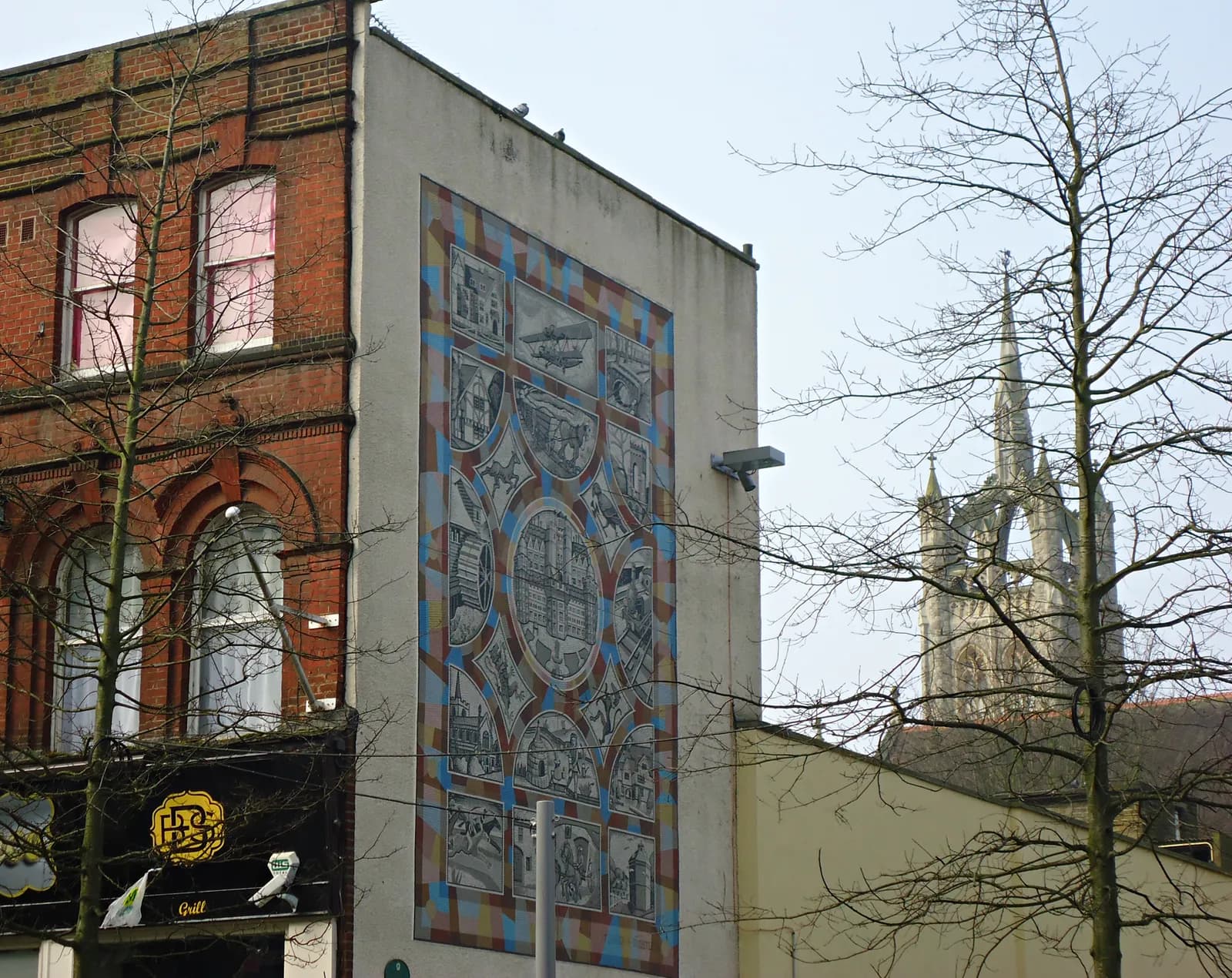 A large mural on the side of a building depicts scenes and a central circular design, next to a brick building and a church steeple in the background, with bare trees in the foreground.