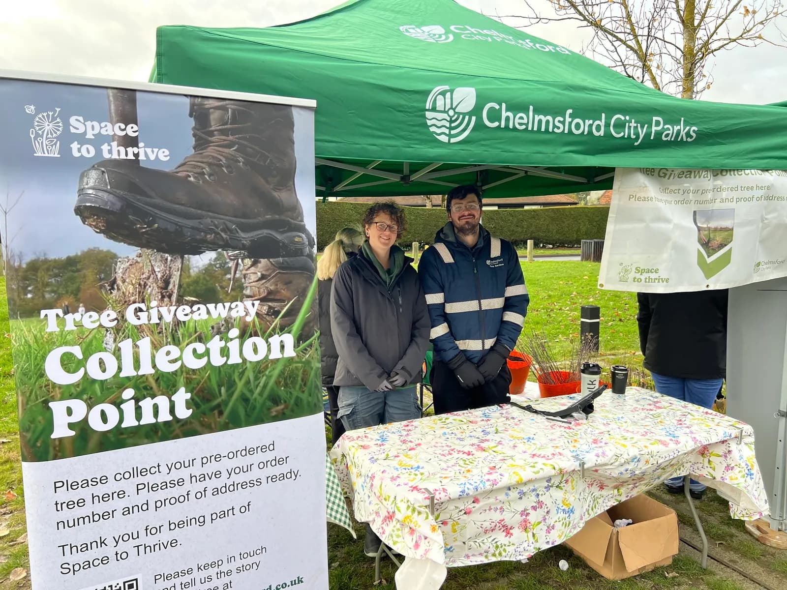 Two people standing behind a table with a floral tablecloth under a green tent, next to a banner for a "Tree Giveaway Collection Point."