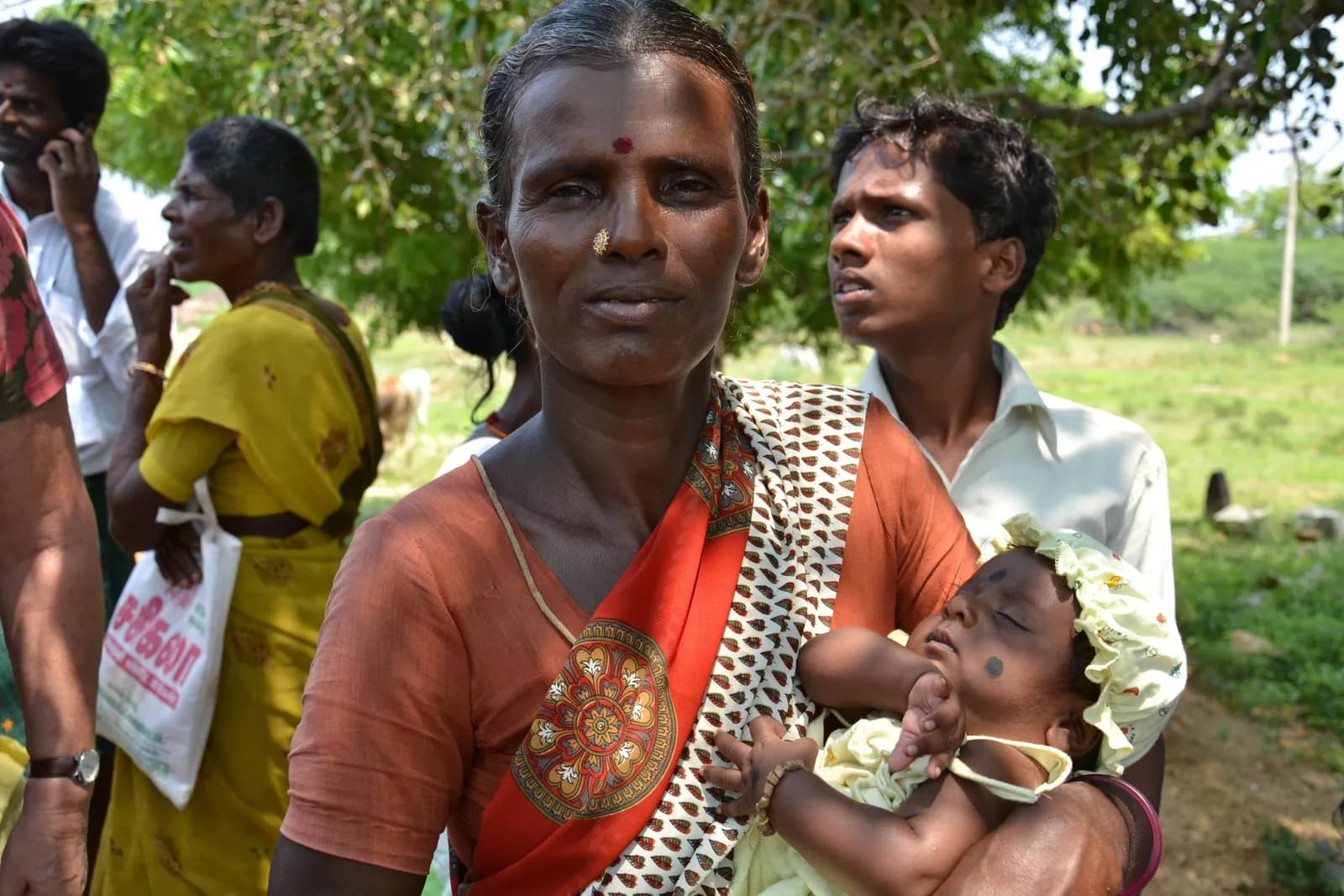 A woman in a red and orange sari holds a sleeping baby, with other people and a green field in the background under a sunny sky.