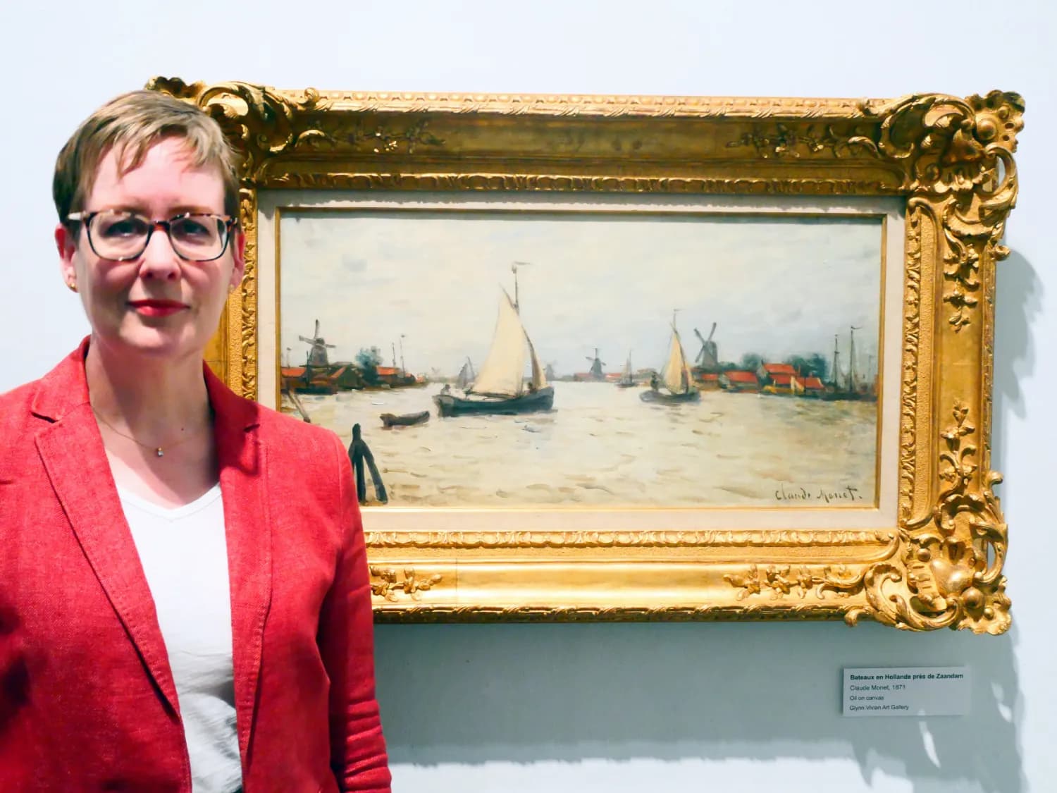A woman in glasses and a red jacket stands next to a framed painting of sailboats on a body of water with windmills and buildings in the background.