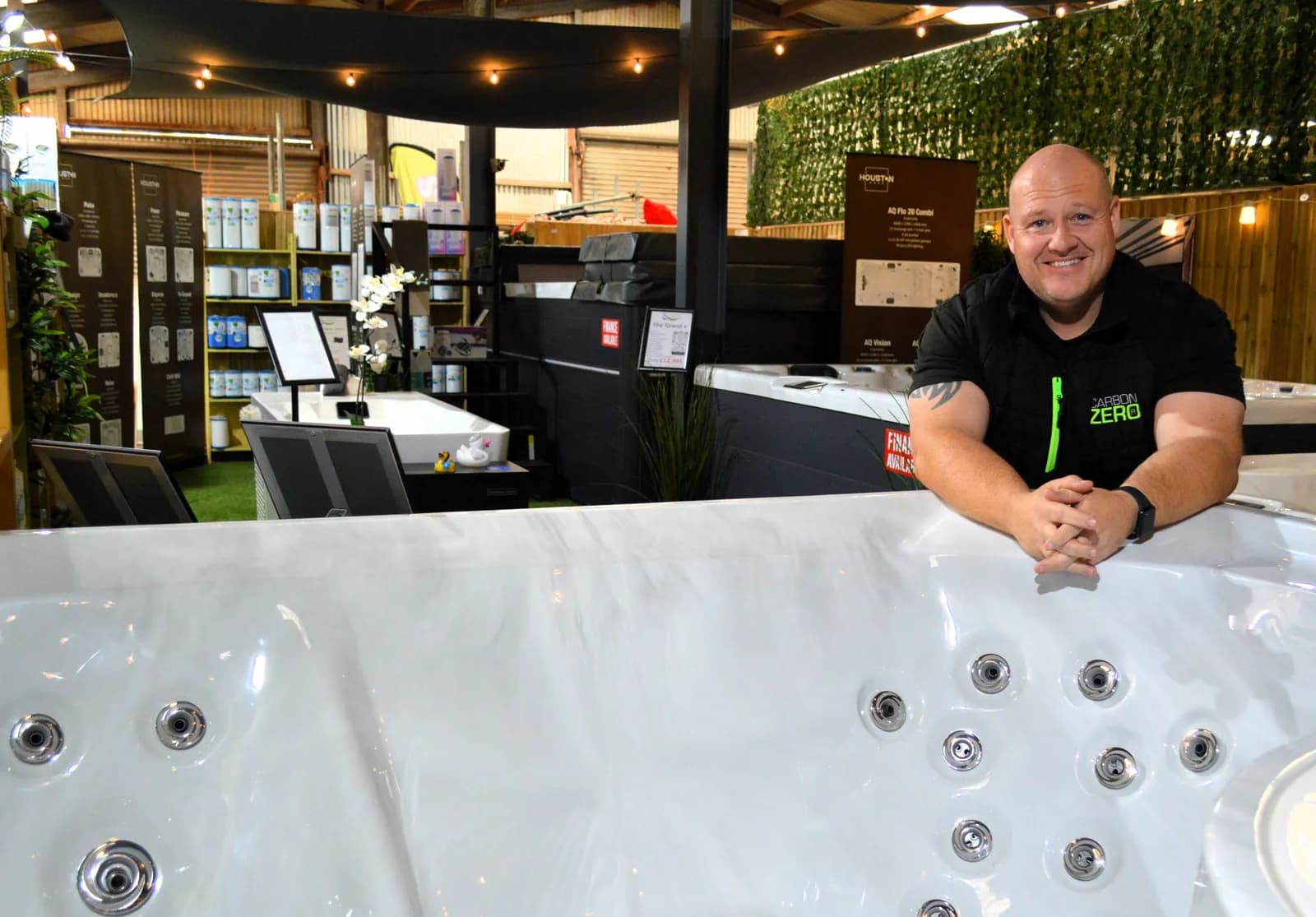 A man in a black polo shirt smiles at the camera, leaning over the edge of a white hot tub, with a retail display of hot tub products behind him.