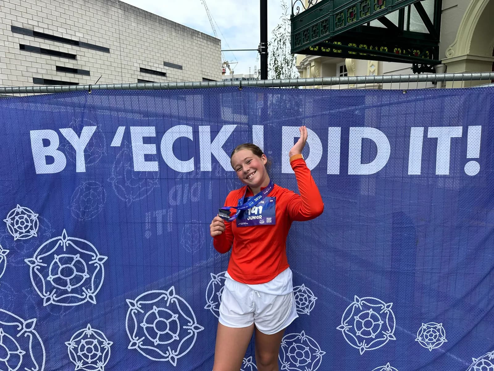 A young girl in an orange shirt and white shorts smiles and holds up a medal in front of a blue banner that reads "BY 'ECK I DID IT!"