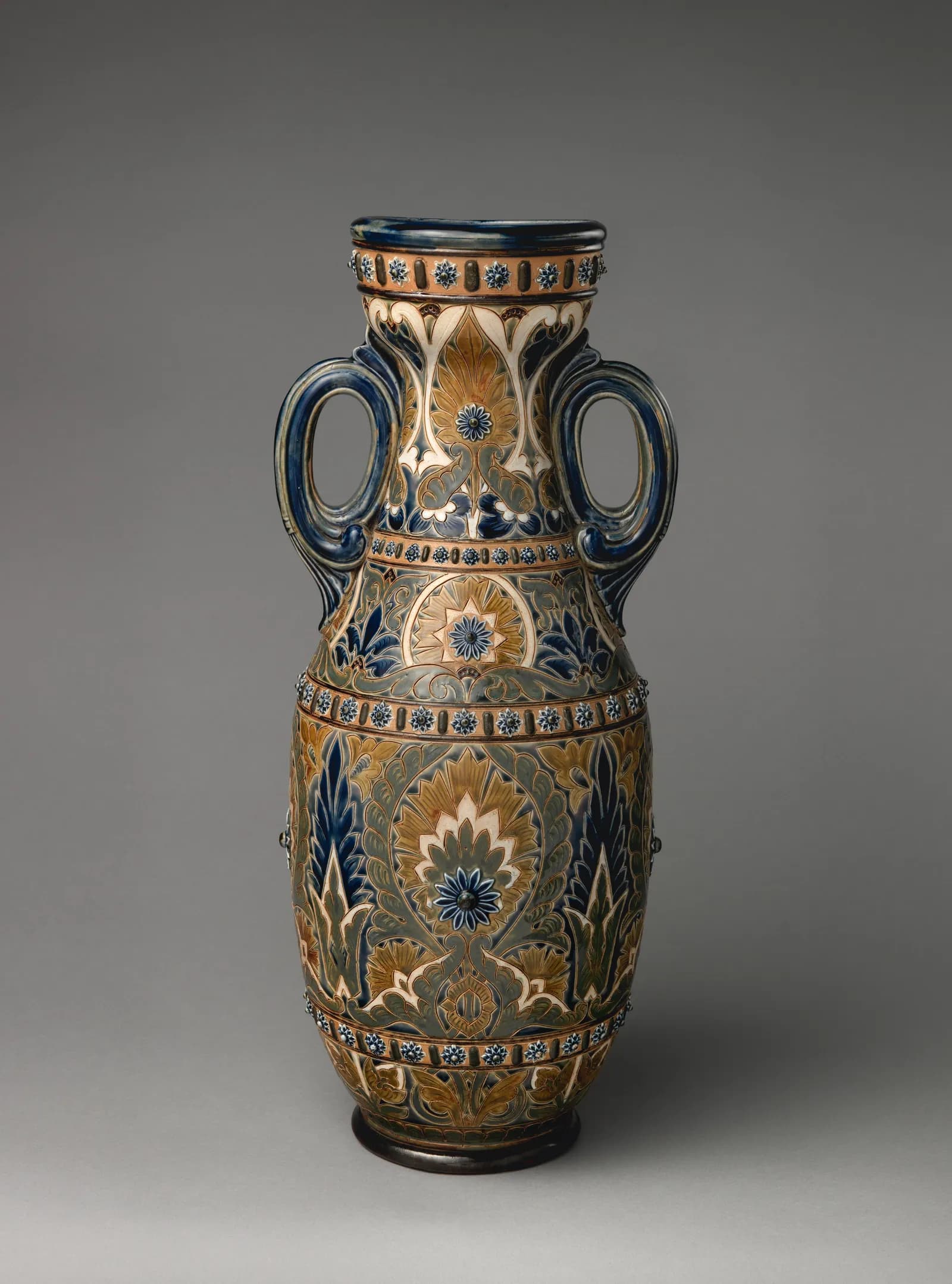 A tall, ornate ceramic vase with two handles, decorated with intricate floral patterns in hues of blue, gold, and white, set against a neutral grey background.