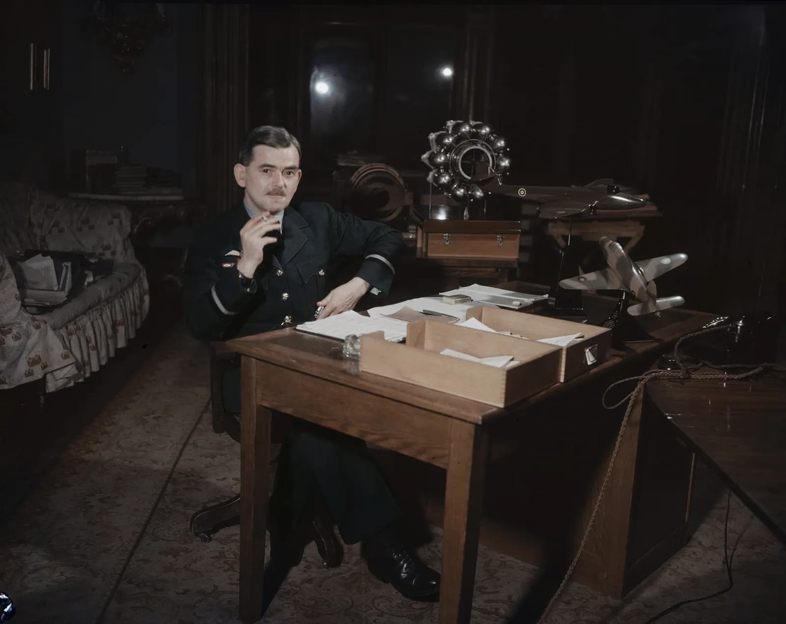 A man in a military uniform with a mustache and slicked-back dark hair sits at a wooden desk with a cigarette between his fingers, with two airplane models and a radial engine on the desk.