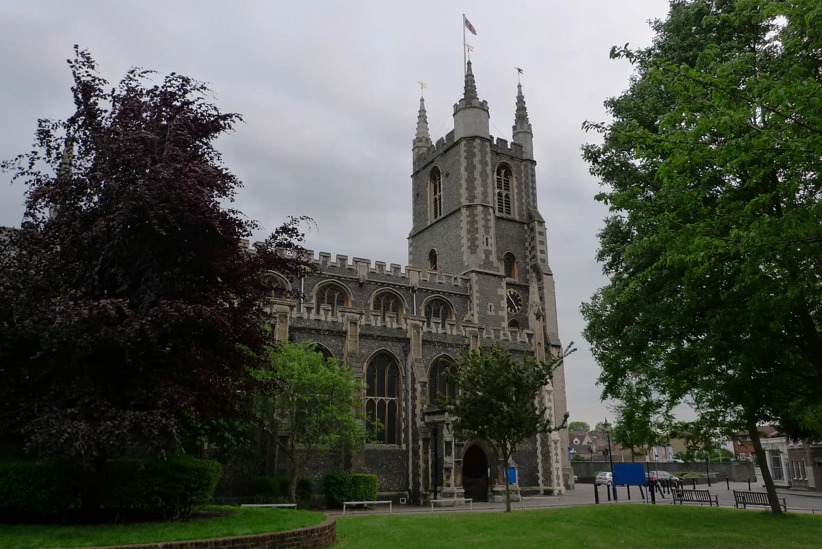A large stone church with a tall tower and a clock, surrounded by green trees and grass under a cloudy sky.