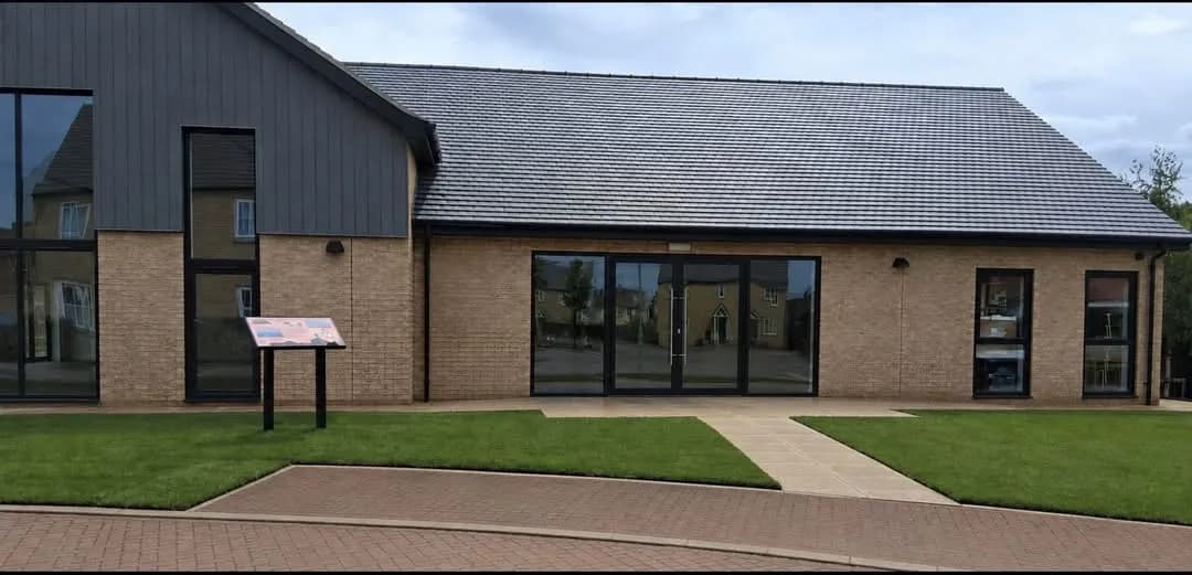 A modern building with a dark grey roof and light brick walls, featuring large glass windows and doors, with green grass and a paved path in front.