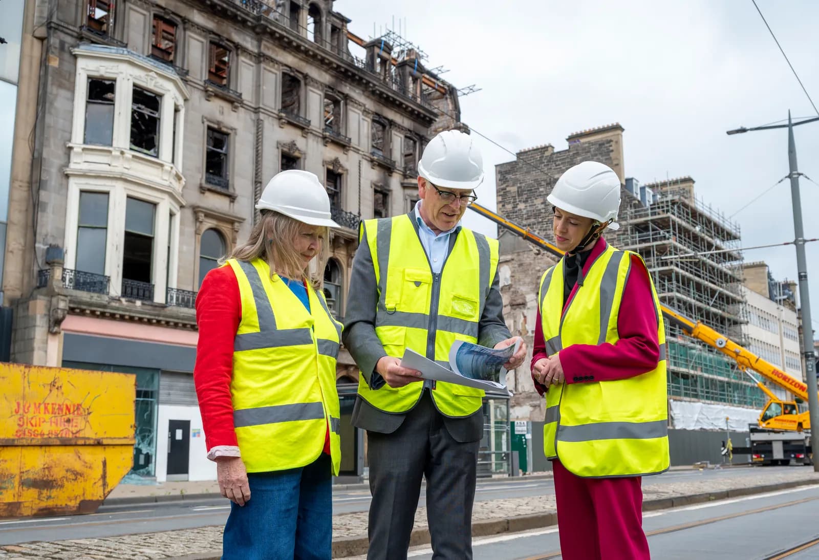 Three people in hard hats and safety vests review documents on a city street with damaged and scaffolding-covered buildings in the background.