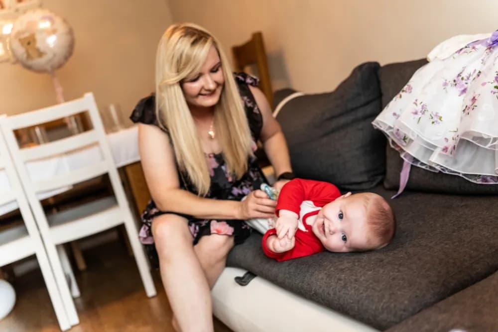A woman with long blonde hair sits on a couch next to a smiling baby wearing a red shirt, who is lying on the couch.