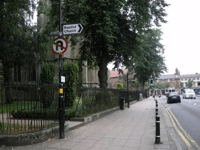 A street scene with a sign pointing to a Baptist Church and a no U-turn sign, with a sidewalk, a black fence, and cars on the road next to buildings in the background.