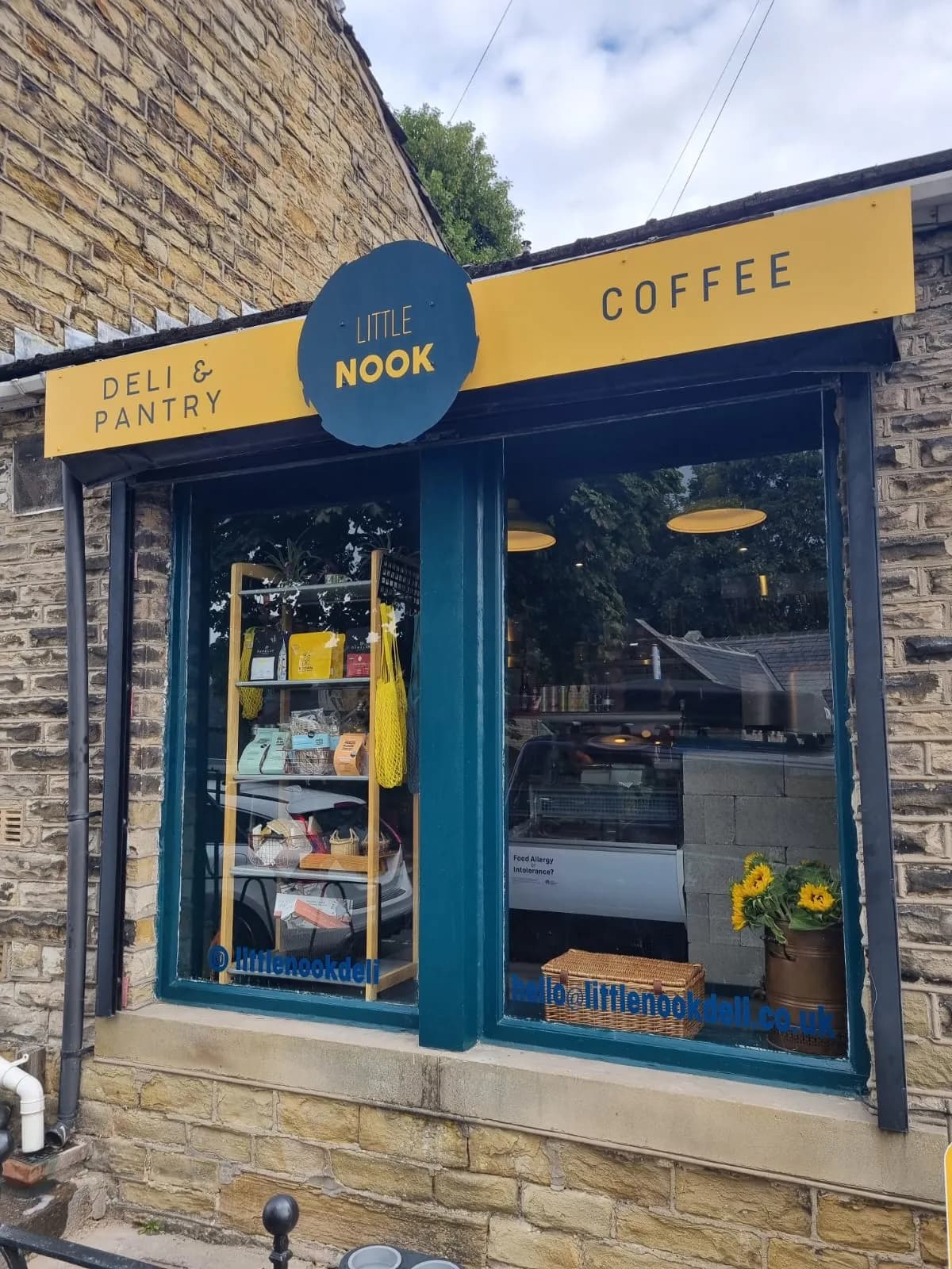 A yellow and blue sign for "Little Nook Deli & Pantry Coffee" above a shop window filled with various deli items and sunflowers.