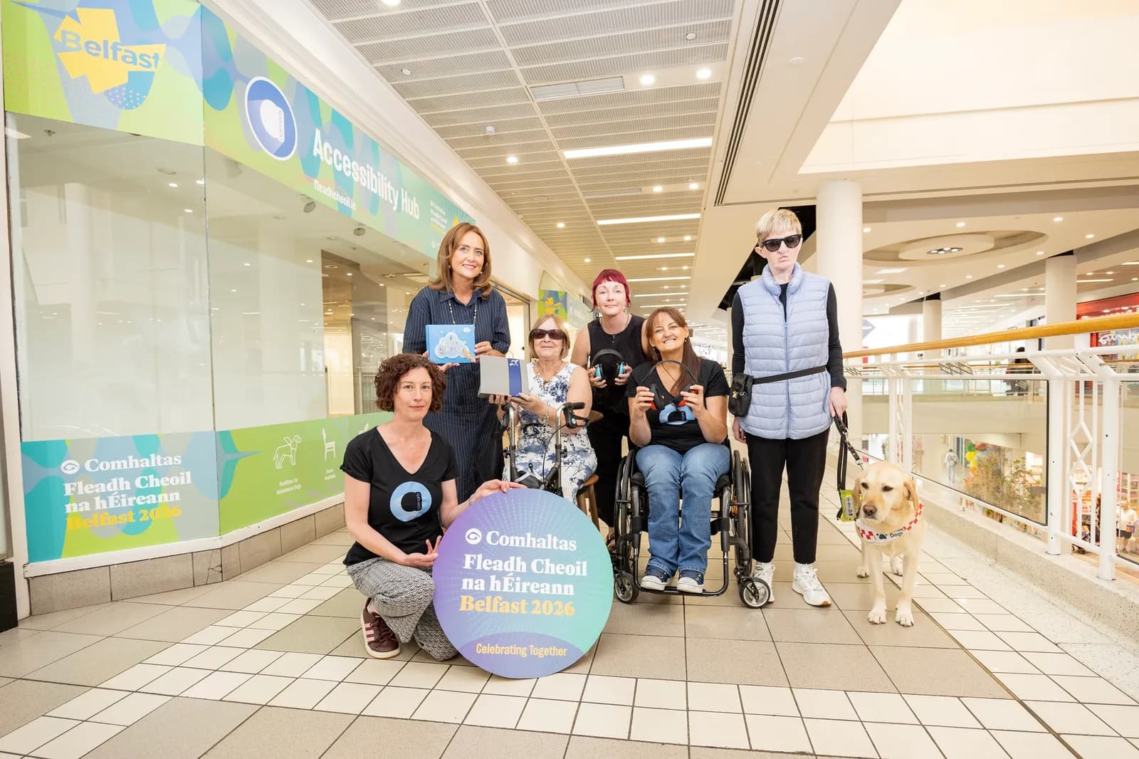 A group of six women, some in wheelchairs and one with a guide dog, stand and pose in a shopping mall with an "Accessibility Hub" sign in the background, holding a circular sign that reads "Comhaltas Fleadh Cheoil na hÉireann Belfast 2026".
