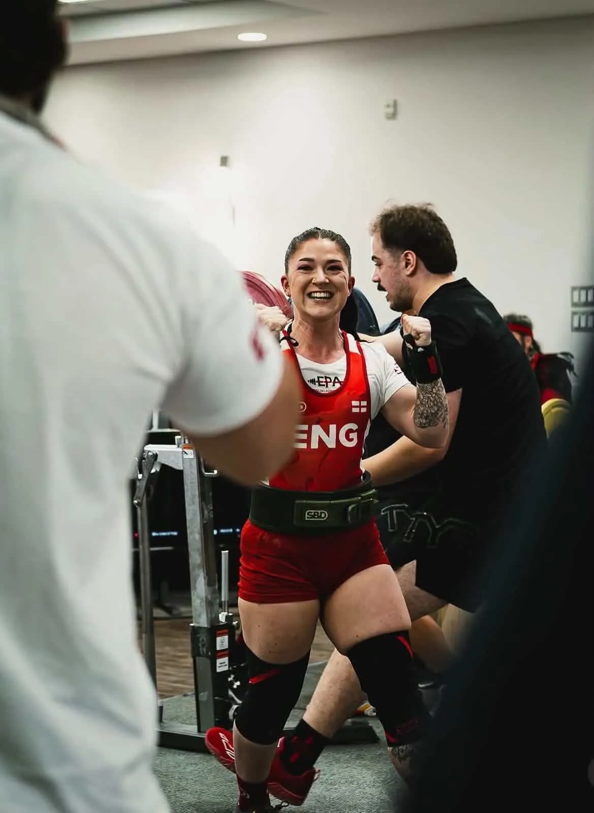 A woman in red shorts and top, with 'ENG' written on it, smiles broadly and flexes her biceps while holding a barbell on her shoulders, surrounded by other people in a gym setting.