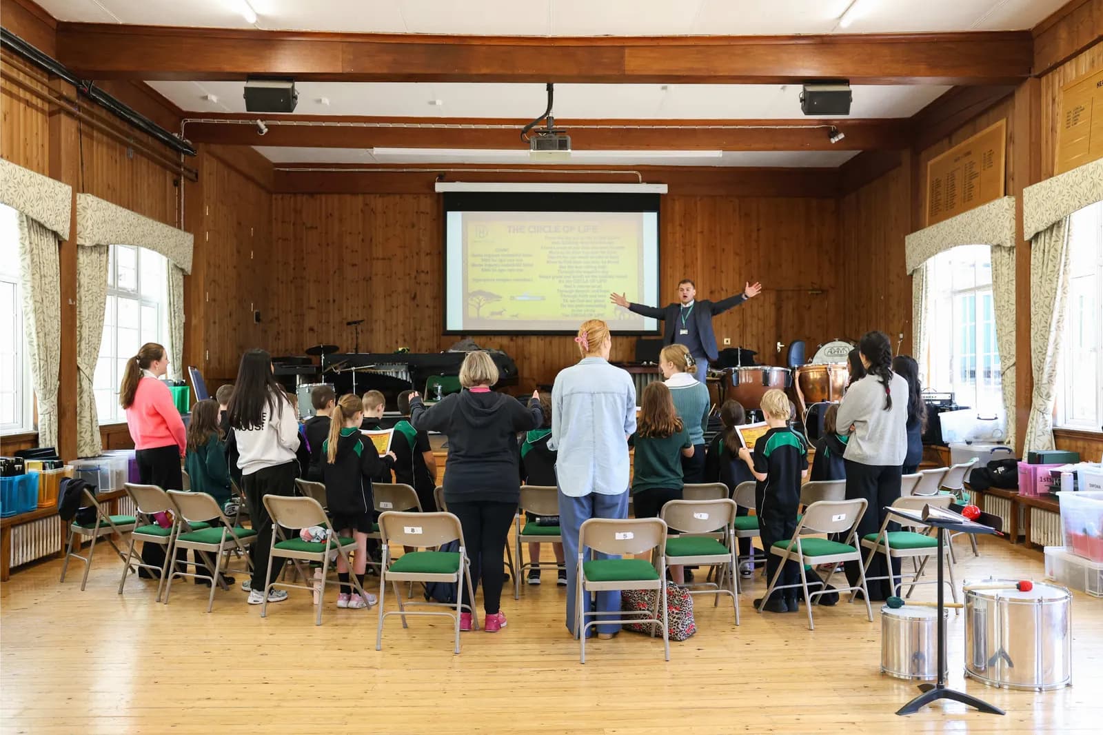 A man with outstretched arms stands on a stage in front of a projector screen, addressing a group of adults and children facing away from the camera in a large room.
