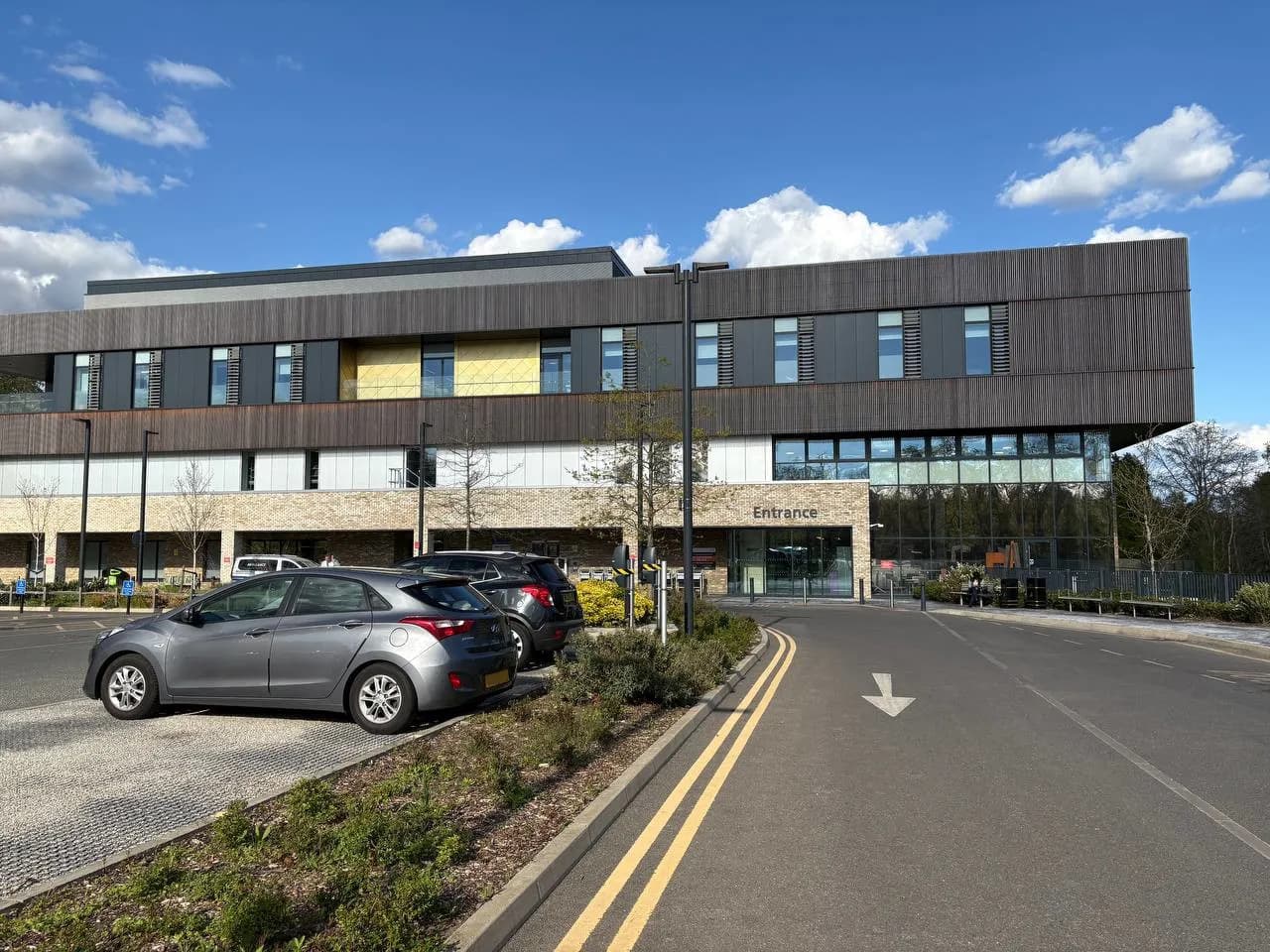 The entrance to a modern hospital building with dark wood siding and large windows, with several cars parked in the foreground under a blue sky.