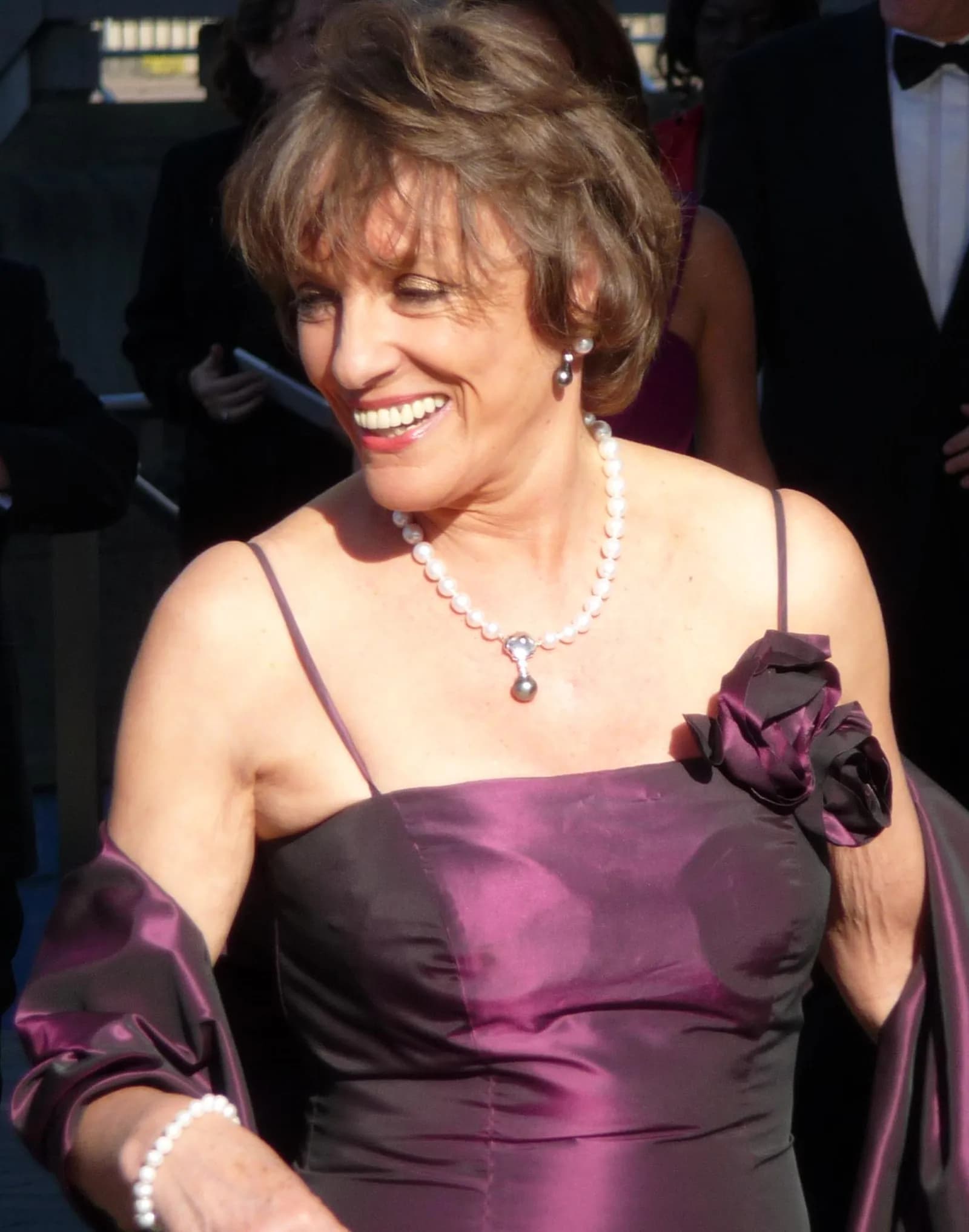 A smiling woman with short brown hair, wearing a dark purple gown and a pearl necklace with a dark pendant, looks towards the left.