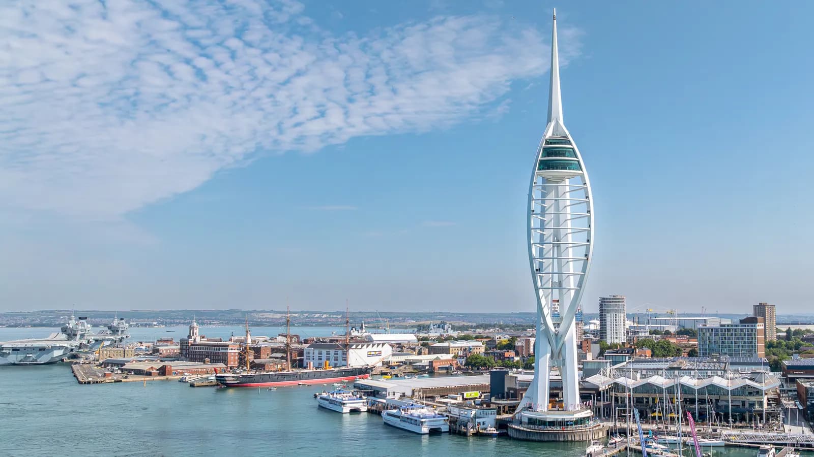 A wide aerial shot of Portsmouth Harbor featuring the iconic Spinnaker Tower on the right, ships docked in the water, and the city buildings in the background under a blue sky.