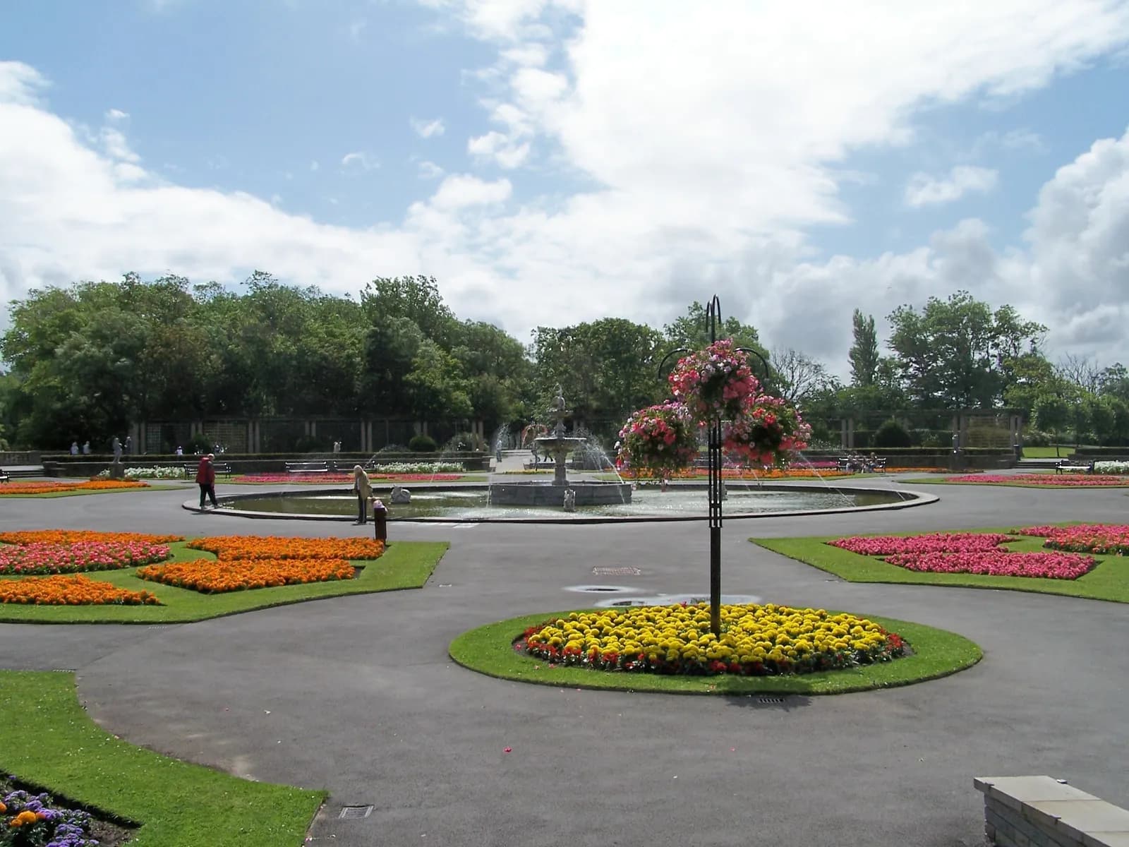 A wide view of a vibrant park with a circular fountain at its center, surrounded by colorful flowerbeds in various shapes, and paved paths with people walking.