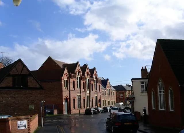 A street scene with traditional brick buildings under a blue sky with white clouds, and parked cars lining the street.