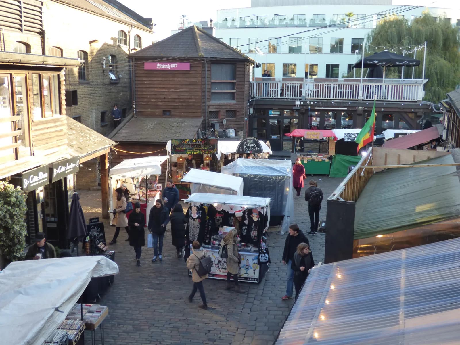 A bustling market scene with many outdoor stalls and people walking on cobblestone ground, surrounded by multi-story buildings.