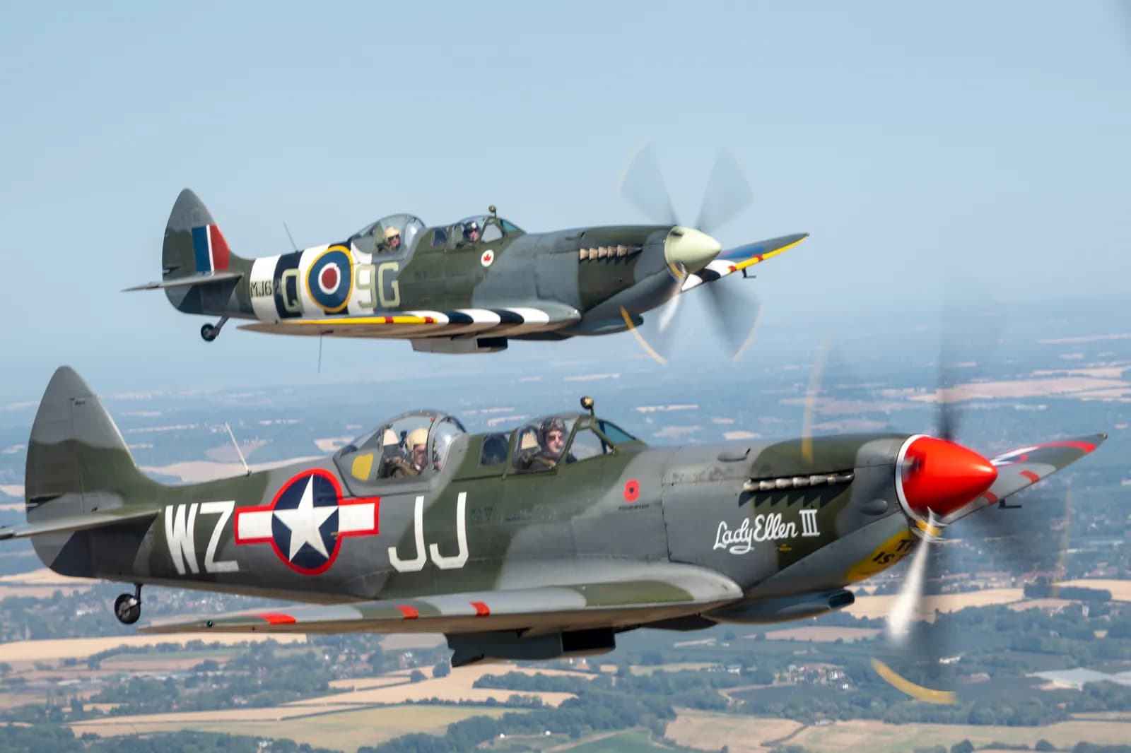 Two vintage military propeller planes, one with a red propeller nose and the other with a white one, fly in formation above a landscape.