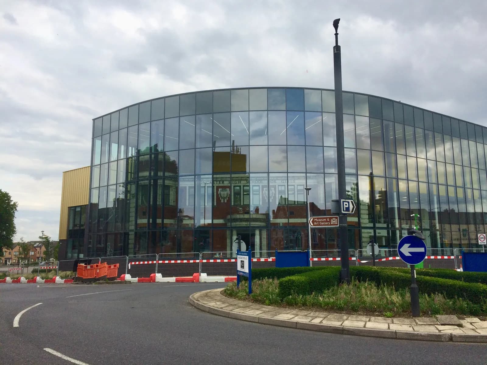 A modern building with a curved glass facade reflects the cloudy sky and surrounding structures, with a road and roundabout in the foreground.