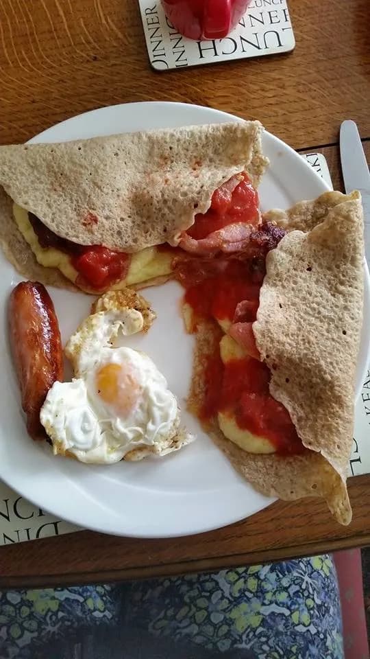 A plate of breakfast food featuring two folded crepes filled with scrambled eggs, bacon, and tomato sauce, alongside a fried egg and a sausage link.