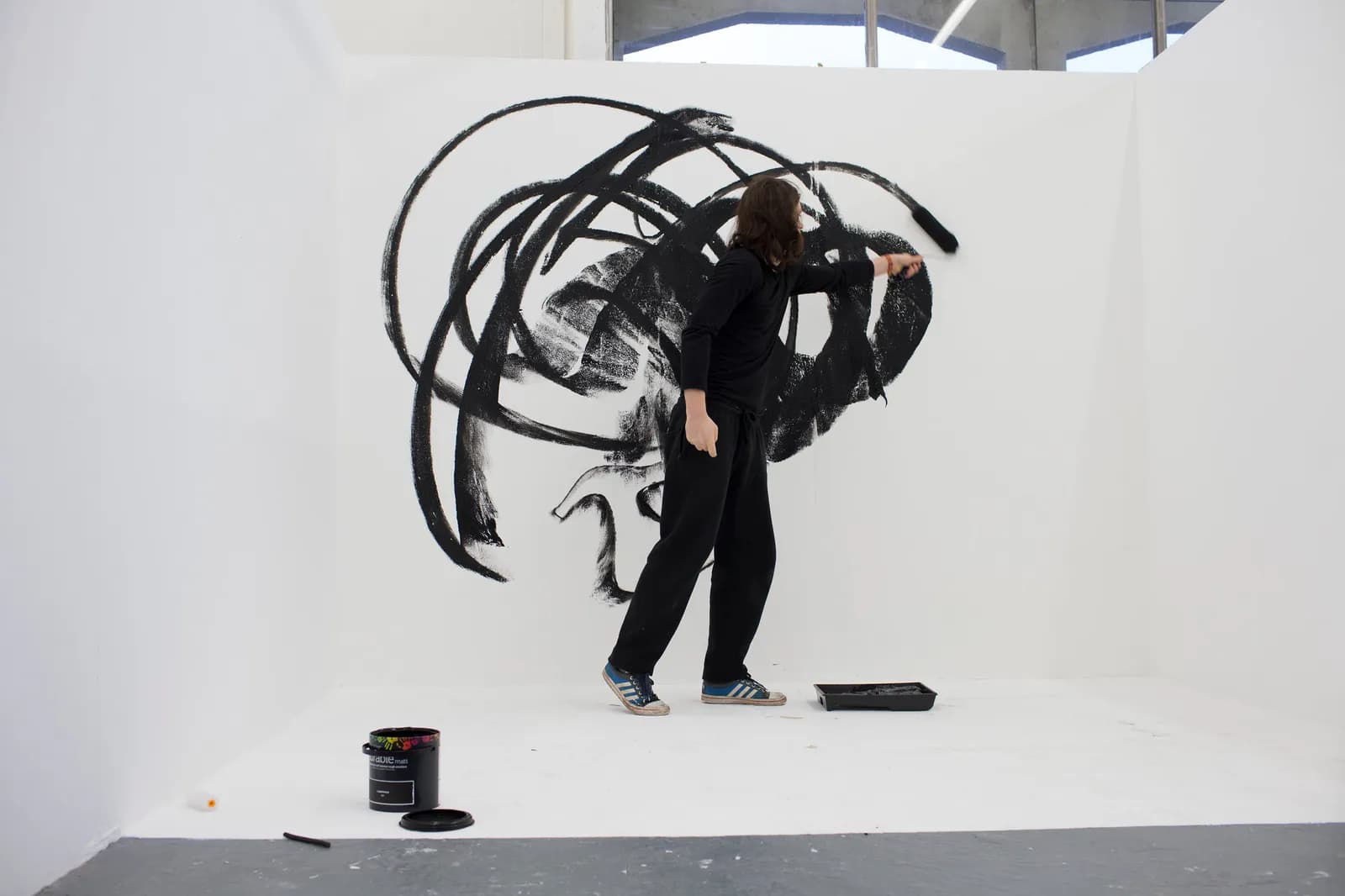 A person in black clothing uses a roller to paint large, sweeping black strokes on a white wall, with a paint can and tray on the floor.