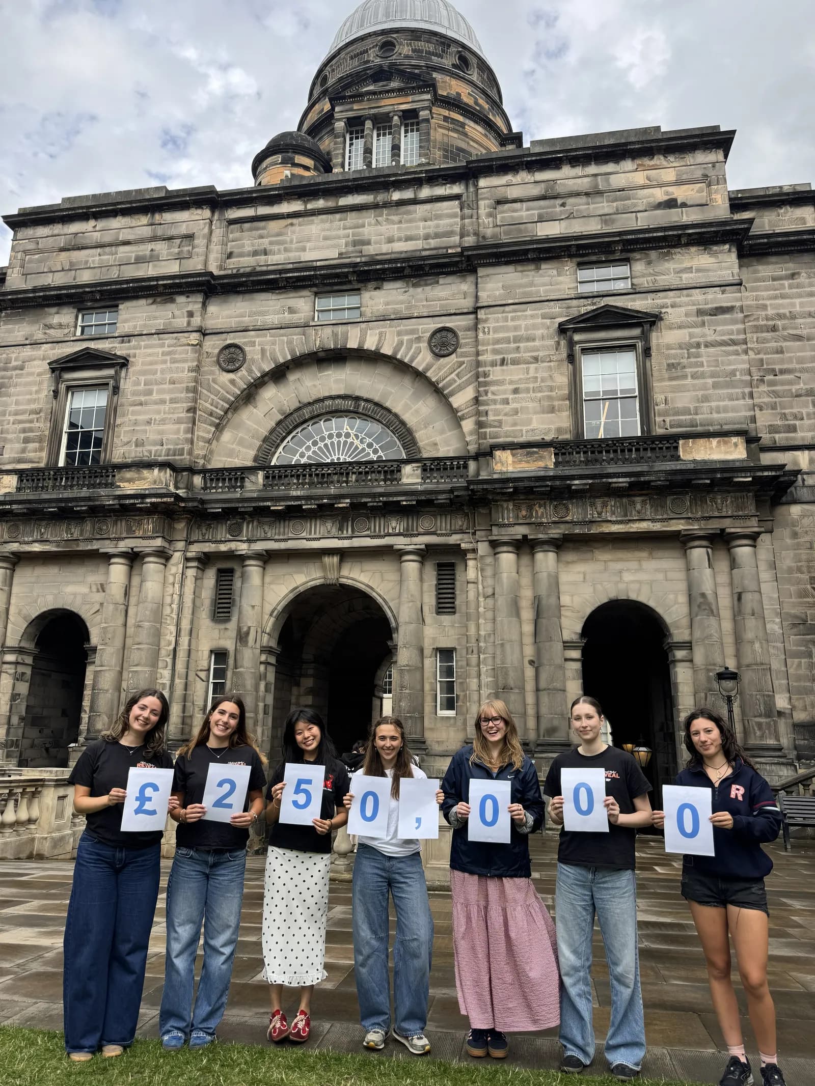 Six young women stand in front of a grand stone building, holding up signs that collectively spell out a large monetary amount: £250,000.