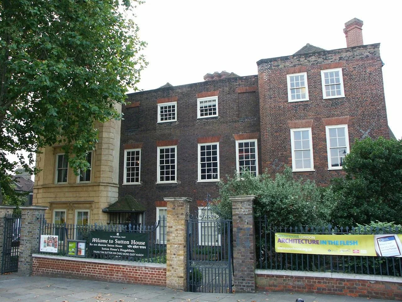 Sutton House, a historic brick building with white-framed windows, stands behind a black metal fence with various informational banners, including one welcoming visitors and another promoting 'Open House London: Architecture in the Flesh'.