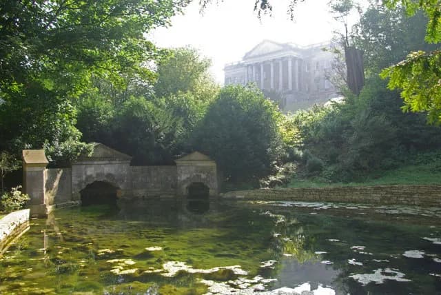 A classical stone building with columns sits atop a hill, partially obscured by green trees, overlooking a serene pond with algae-covered water and stone arches.