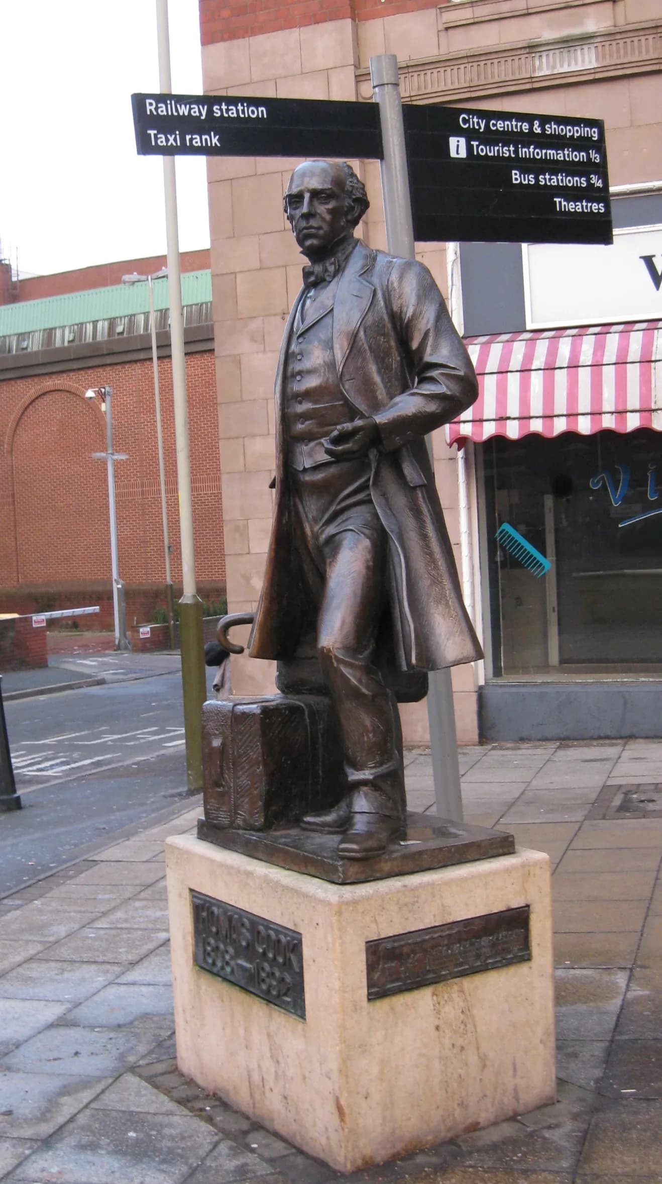 A bronze statue of a man in a formal suit and long coat stands on a stone pedestal on a city sidewalk, with street signs pointing to "Railway station" and "City centre & shopping" behind him.