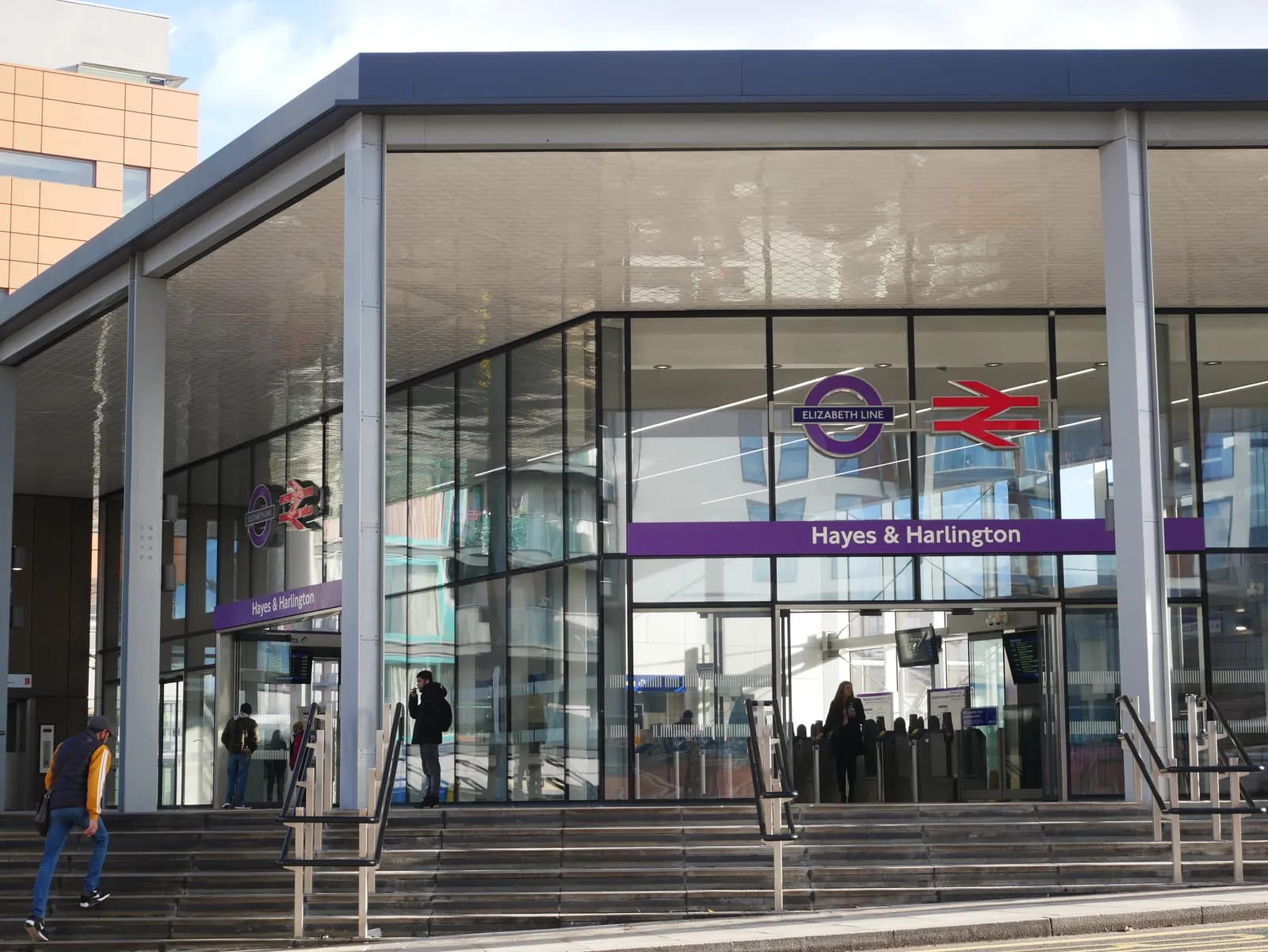 The entrance to the Hayes & Harlington Elizabeth Line station, with its modern glass and steel facade, purple signage, and steps leading up to the turnstiles where people are entering.