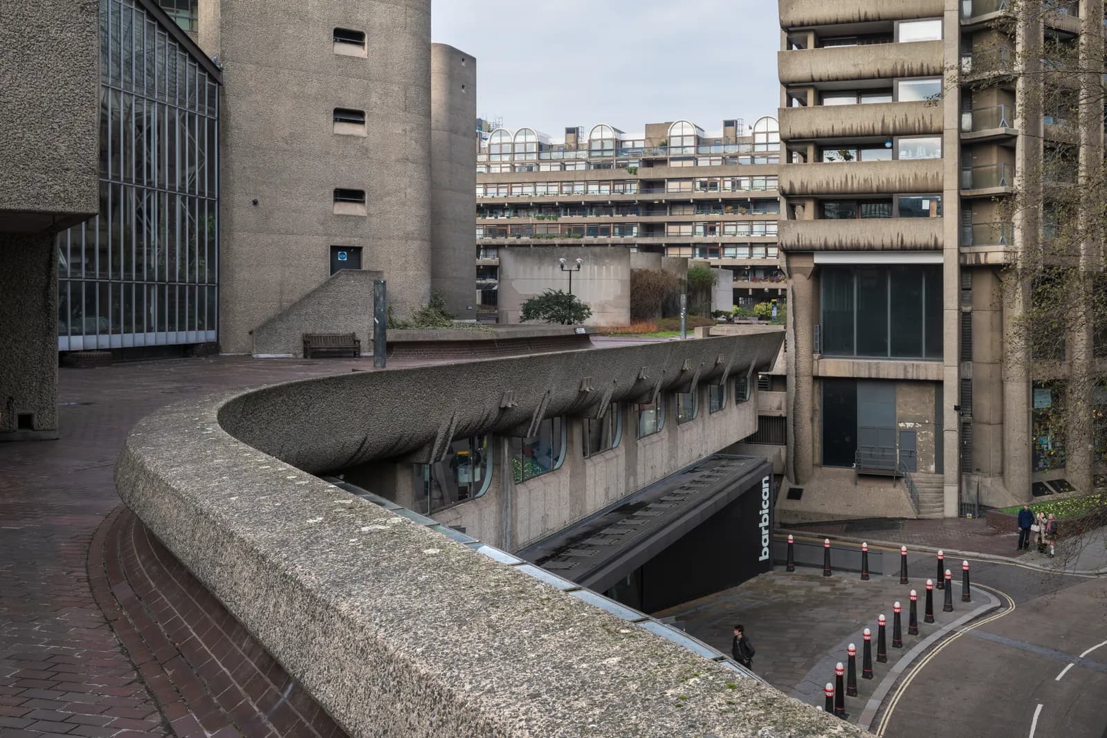 A sweeping concrete walkway in foreground, curving towards the right, overlooks a brutalist architectural complex with gray buildings, multiple levels, and an underpass with the word 'barbican' visible, all under an overcast sky.