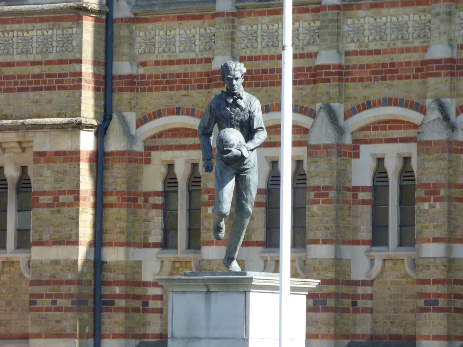 A bronze statue of a man holding a ball stands in front of a brick building with decorative carvings, under clear skies.