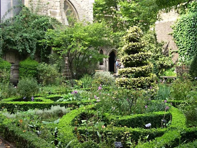 A lush, green garden with manicured hedges and various plants, featuring an evergreen topiary tree in the center and a building with archways in the background.