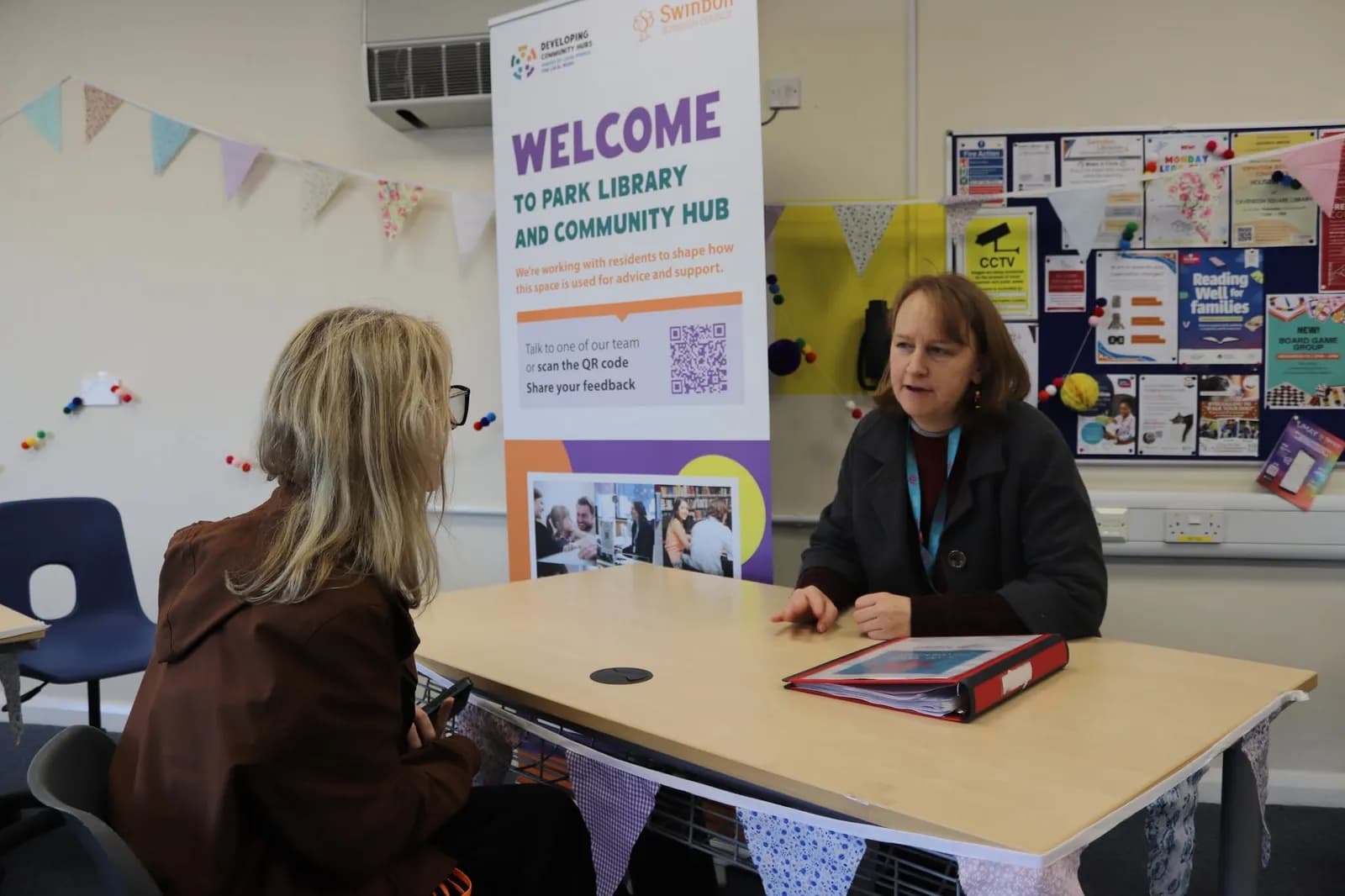 A woman with light brown hair sits at a desk across from another person, engaged in conversation, with a large 'Welcome to Park Library and Community Hub' banner behind her.