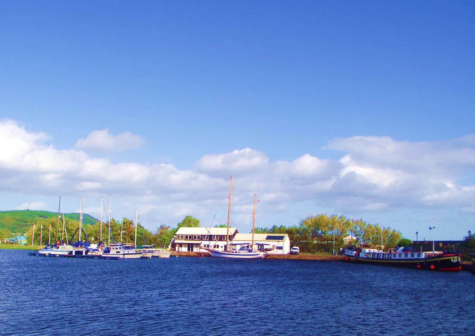 A picturesque marina with several boats docked along a brown shore, a white building, and lush green hills under a bright blue sky with fluffy white clouds.