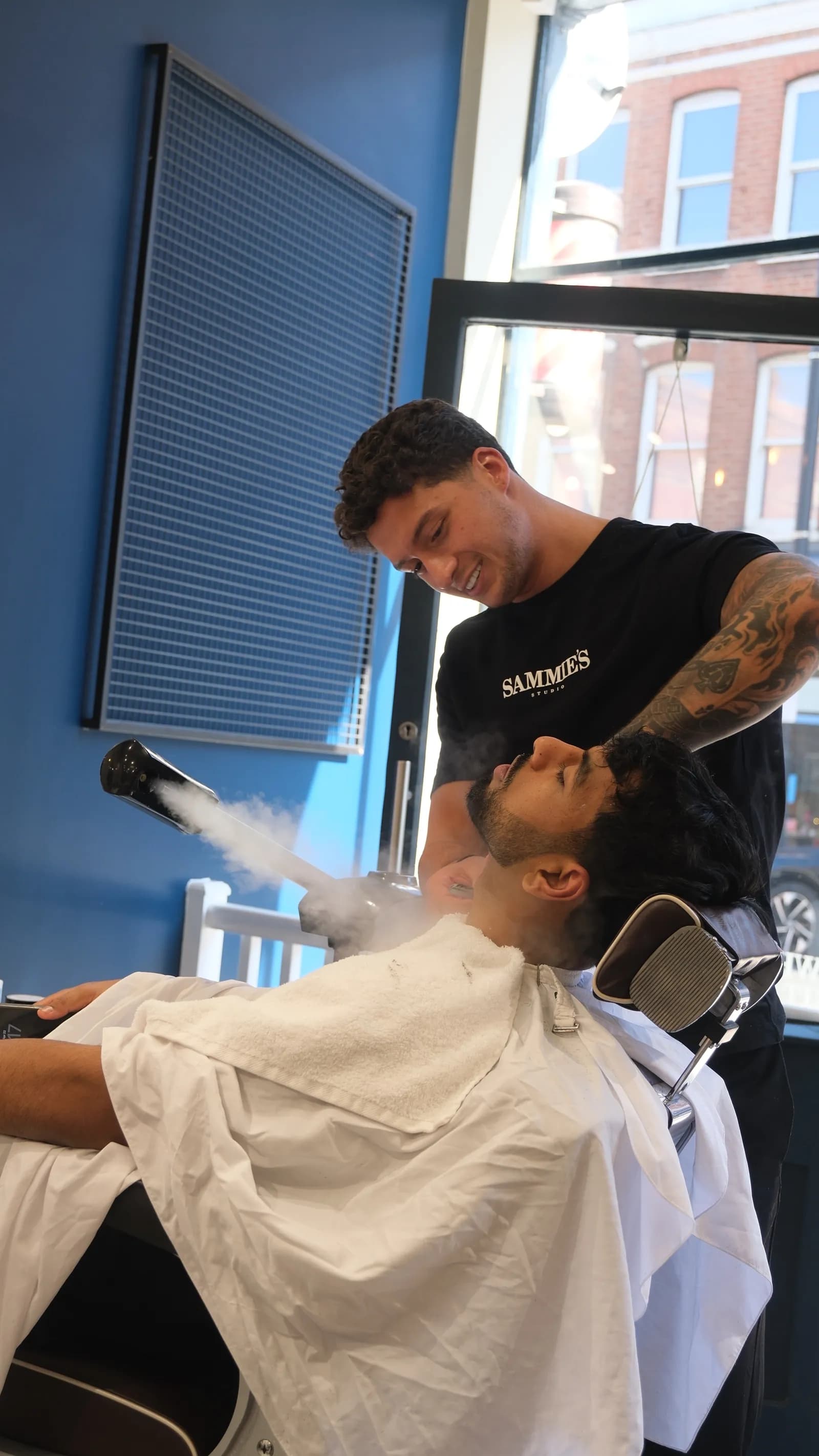 A barber applies steam to a client's face who is sitting in a barber chair covered with a white cape.