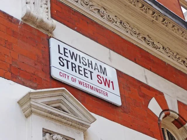 A white street sign with black and red text for "LEWISHAM STREET SW1" and "CITY OF WESTMINSTER" mounted on a brick wall with white architectural details.