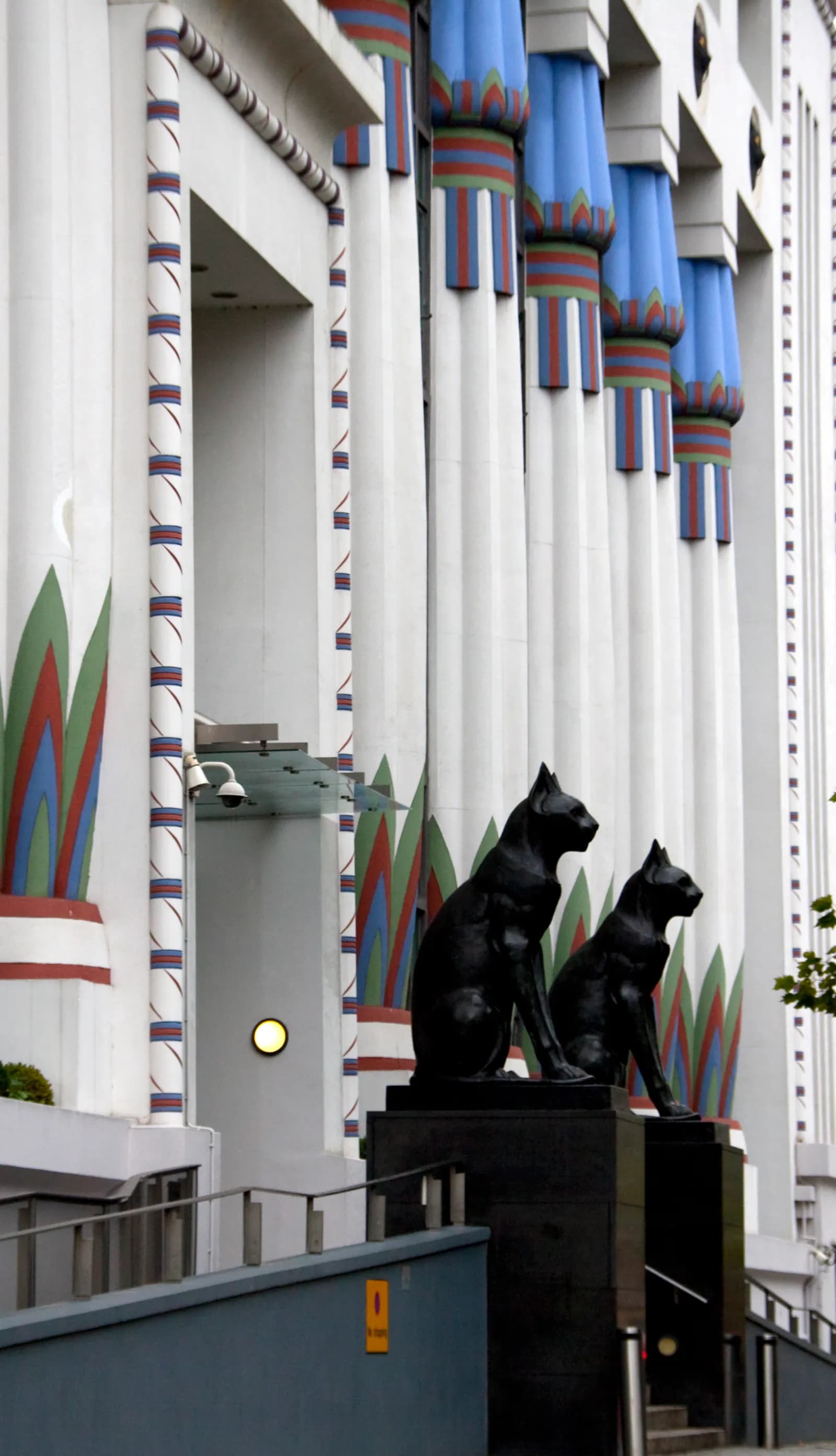 Two large black cat statues sit proudly at the base of a building with blue, red, and green Egyptian-inspired columns.