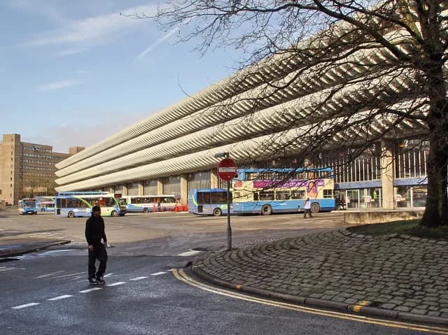 A large concrete building with a ribbed facade serves as a bus station, with several buses parked and a person walking in the foreground.